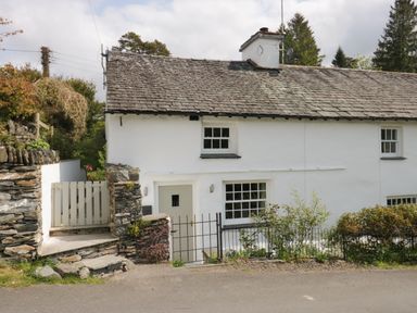 A white stone cottage with a tiled roof and small garden behind a metal fence at Old Farm Cottage in Skelwith Fold