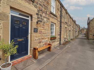 A stone row house with a navy door number 2 a wooden bench and a potted plant outside on a street at Lark Rise Alnmouth