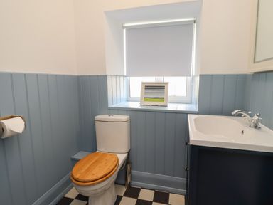 A bathroom with a toilet that has a wooden seat a sink with a faucet a window with a white roller blind and a framed picture on the windowsill at Lark Rise in Alnmouth