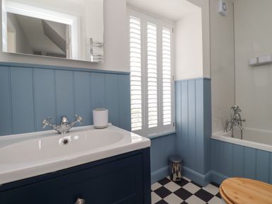 A bathroom with a sink, bathtub, toilet with wooden seat, window with white shutters, and blue paneled walls at Lark Rise Alnmouth