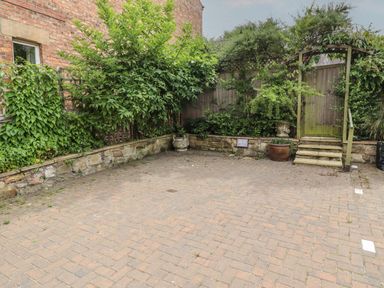A paved courtyard with a wooden arched gate steps and green plants near a brick building at Lark Rise in Alnmouth