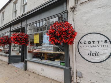 The exterior of a deli store with hanging baskets of red flowers at Scott's of Alnmouth