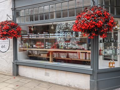 A deli shop window with jars and bottles on shelves and red flowers hanging outside at Lark Rise in Alnmouth