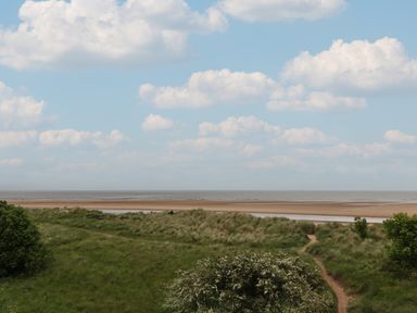 A sandy beach with grass dunes and a path leading to the shore under a cloudy sky at Lark Rise in Alnmouth