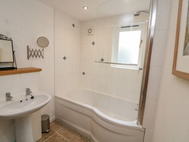 A bathroom with a bathtub and shower, a pedestal sink, a small wall-mounted mirror and a window at The Loft Cottage in Totnes
