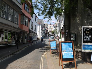A street with shops and cafes along the sides with signs and people walking at The Loft Cottage in Totnes