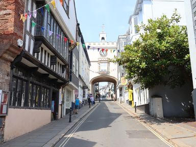 A street with people walking and buildings on both sides with a clock tower archway at the end and colorful flags hanging above at The Loft Cottage in Totnes