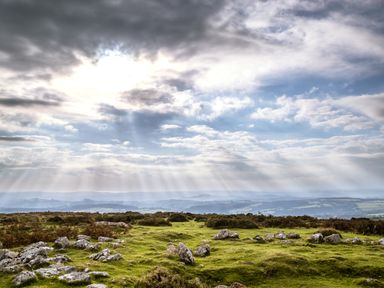 A grassy landscape with scattered rocks under a cloudy sky with sun rays at The Loft Cottage in Totnes
