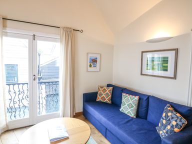 A living room with a blue sofa and patterned cushions near glass doors with curtains at The Loft Cottage in Totnes