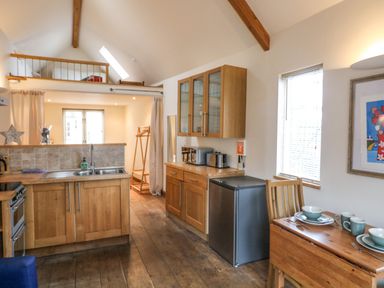 A kitchen with wooden cabinets a small refrigerator a dining table set with dishes and a loft area above at The Loft Cottage in Totnes