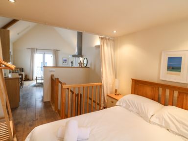 A bedroom with a wooden bed and white bedding next to a wooden railing and an open view to a living area at The Loft Cottage in Totnes