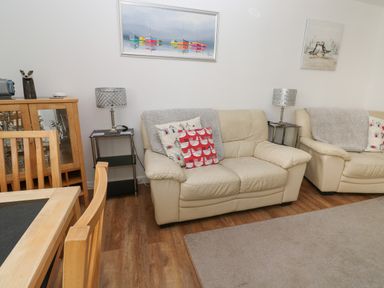 A living room with two beige sofas, cushions, side tables with lamps, a wooden dining table with chairs, and wall paintings at Badger Sett in Granite Henge near Looe