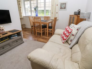 A living room with a beige sofa and colorful cushions a wooden dining table with chairs and a TV on a stand at Badger Sett in Granite Henge near Looe