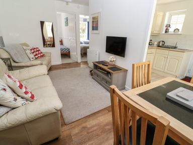 A living room with beige sofas a wall mounted TV a wooden dining table and chairs and a kitchen visible through a doorway at Badger Sett in Granite Henge near Looe