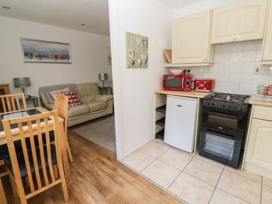 A kitchen with a black oven red microwave and toaster next to a small white fridge and a dining area with wooden chairs and table and a living room with a beige sofa at Badger Sett in Granite Henge near Looe