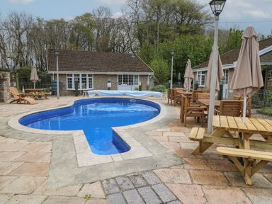 An outdoor pool area with wooden tables chairs umbrellas and cottages in the background at Badger Sett in Granite Henge near Looe