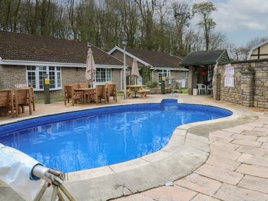 An outdoor swimming pool with blue water surrounded by stone pavement and wooden tables with umbrellas at Badger Sett in Granite Henge near Looe