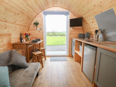 A small wooden kitchen and dining area with a sofa a table with stools kitchen appliances and an open door showing a grassy outside view at Nebo Pod in Amlwch
