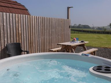 An outdoor area with a hot tub in the foreground a wooden picnic table with tableware and chairs next to a wooden fence at Nebo Pod in Amlwch