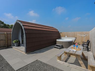 An outdoor area with a wooden pod building a hot tub a picnic table with glasses and a chair at Nebo Pod in Amlwch