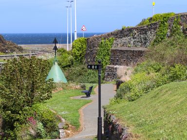 A path with a bench and signpost next to stone walls and greenery by the sea at Nebo Pod in Amlwch