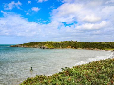 A coastal bay with a rocky shoreline and green vegetation under a partly cloudy sky at Nebo Pod in Amlwch