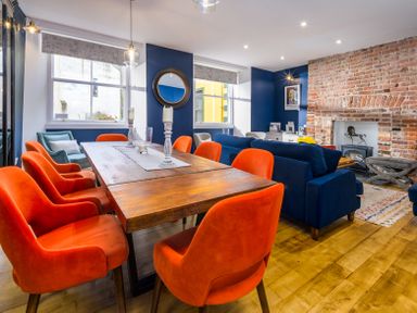 A dining area with orange chairs around a wooden table next to a living room with blue sofas and a brick fireplace at The Indigo House in Berwick-Upon-Tweed