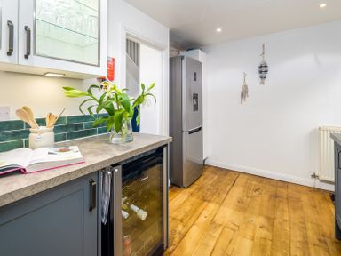 A kitchen with a countertop holding a vase with flowers an open book a utensil holder and a wine cooler next to a refrigerator and wooden flooring at The Indigo House in Berwick-Upon-Tweed