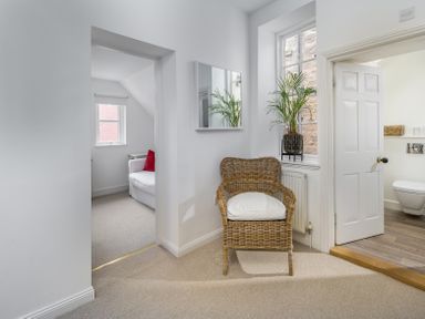 A wicker chair with a white cushion under a window with a plant next to a doorway leading to a bathroom and another room with a white sofa at The Indigo House in Berwick-Upon-Tweed