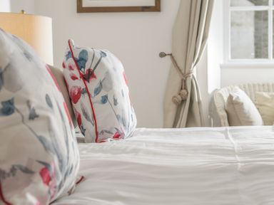 A close up of floral patterned pillows on a bed next to a lamp and a window with curtains at The Indigo House in Berwick-Upon-Tweed