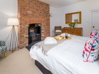 A bedroom with a brick fireplace a bed with white linens and floral pillows a wooden dresser and a floor lamp at The Indigo House in Berwick-Upon-Tweed
