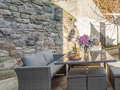 An outdoor patio with cushioned wicker seating a glass table with flowers and food against a stone wall at The Indigo House in Berwick-Upon-Tweed