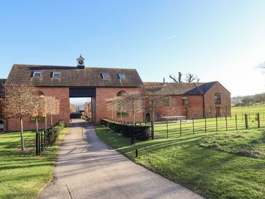 A paved driveway leading to a brick building with a clock tower and fenced lawns at The Millhouse in Ledbury near Welland