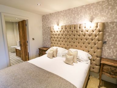 A bedroom with a bed with beige tufted headboard and four pillows two wooden bedside tables and an open door to a bathroom at The Millhouse in Ledbury near Welland