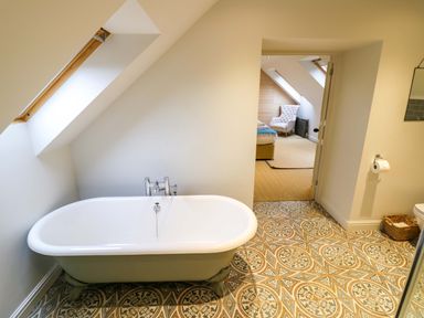 A bathroom with a bathtub and patterned floor tiles leading to a bedroom with a chair at The Millhouse Ledbury near Welland