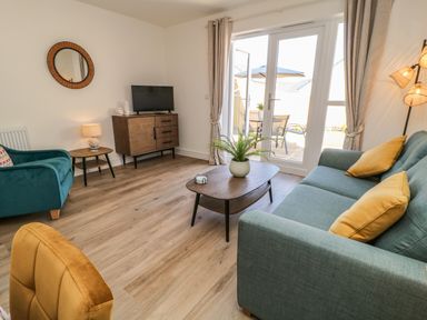 A living room with a teal sofa and yellow cushions a green armchair a wooden coffee table with a plant a cabinet with a television and patio doors with curtains at Pebble Bay in Appledore