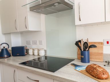 A kitchen countertop with a blue kettle and toaster a stovetop a wooden cutting board a blue utensil holder with cooking utensils and a loaf of bread on a cutting board at Pebble Bay in Appledore
