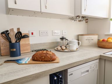 A kitchen counter with a loaf of bread on a cutting board utensils in a holder a teapot and creamer on a tray a bread bin and a fruit basket at Pebble Bay in Appledore