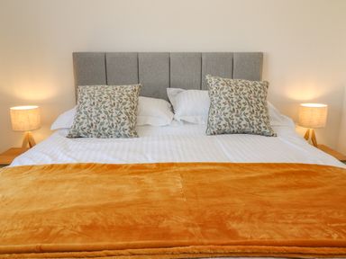 A bed with a grey headboard two patterned pillows white sheets an orange blanket and two bedside lamps at Pebble Bay in Appledore