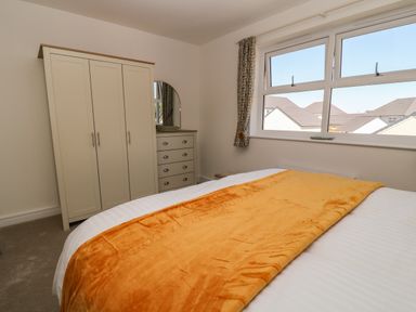 A bedroom with a bed covered with a white sheet and an orange blanket a wardrobe and a chest of drawers with a mirror and a window with curtains at Pebble Bay in Appledore