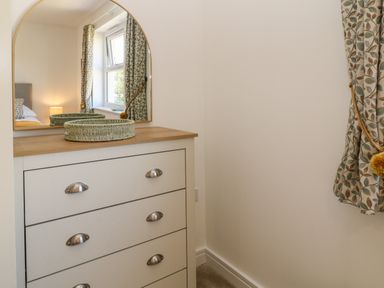 A chest of drawers with a mirror and basket beside a window with patterned curtains at Pebble Bay in Appledore