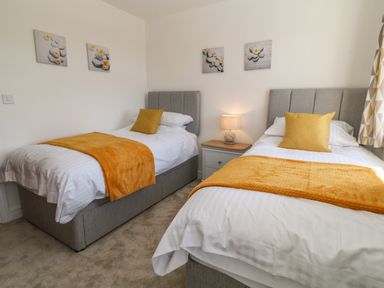 A bedroom with two single beds with yellow pillows and throws a bedside table with lamp and floral wall art at Pebble Bay in Appledore