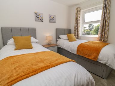 A bedroom with two single beds with grey headboards orange blankets and pillows a bedside table with a lamp and a window with patterned curtains at Pebble Bay in Appledore
