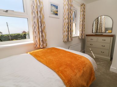 A bedroom with a bed covered by an orange throw a dresser with a mirror and patterned curtains at Pebble Bay in Appledore