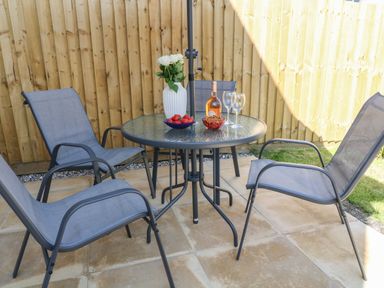 A patio set with four chairs and a round glass table with a vase of white flowers a bottle of wine two wine glasses and bowls of strawberries and snacks at Pebble Bay in Appledore