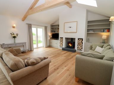 A living room with two sofas a fireplace wood logs shelves and a glass door at Cotswold View in Shipton Under Wychwood