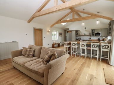 An open plan living room and kitchen with a beige sofa wooden beams and bar stools at Cotswold View in Shipton Under Wychwood