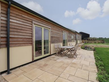 A patio with a wooden table and chairs outside a building with wooden siding at Cotswold View in Shipton Under Wychwood