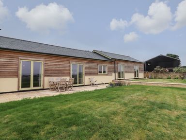A wooden house with multiple glass doors and a patio with a wooden table and chairs in a grassy yard at Cotswold View in Shipton Under Wychwood