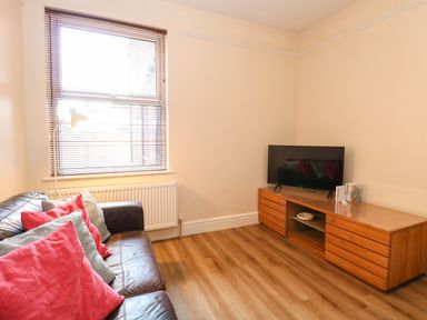 A living room with a brown leather sofa with red and gray cushions a wooden TV stand with a television and a window with wooden blinds at Seaside House in Gorleston-On-Sea